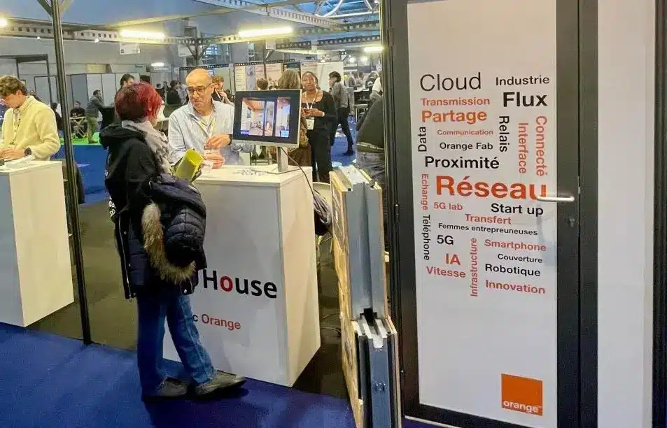 Stand de UHouse sur le salon de l'industrie du futur à Mulhouse. On y voir à droite une affiche de Orange l'opérateur télécom français, et à sa gauche le stand d'UHouse composé d'un écran et de supports papiers sur un comptoir. Derrière Hamid Chibane le dirigeant de la société et devant une dame en pleine discussion de découverte du concept.